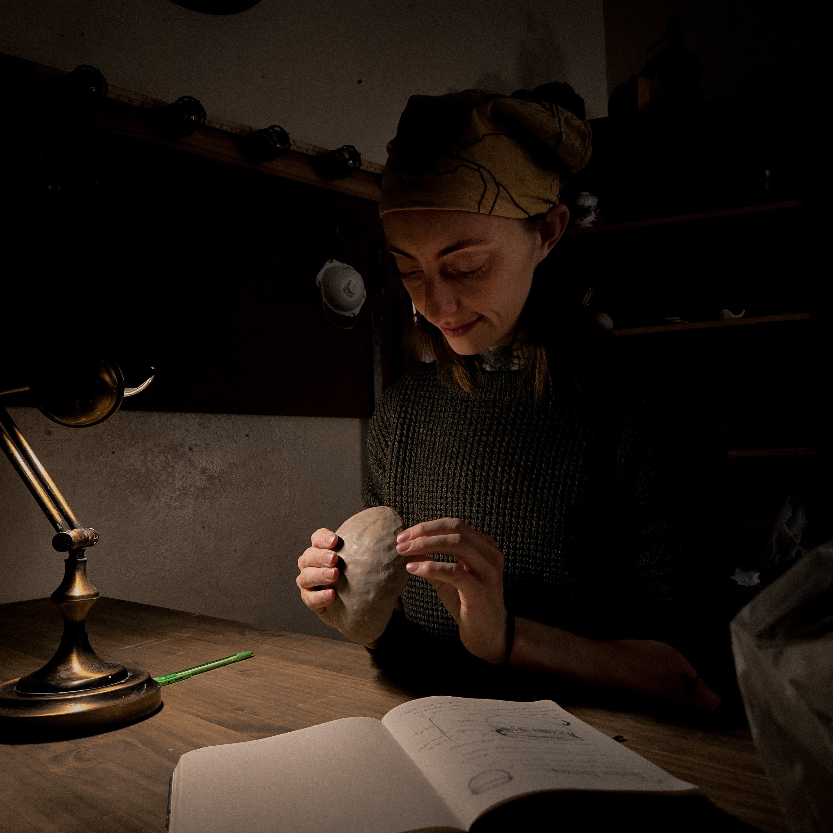 Person pinching clay to form a pinch pot in a dimly lit room with a lamp and sketchbook.